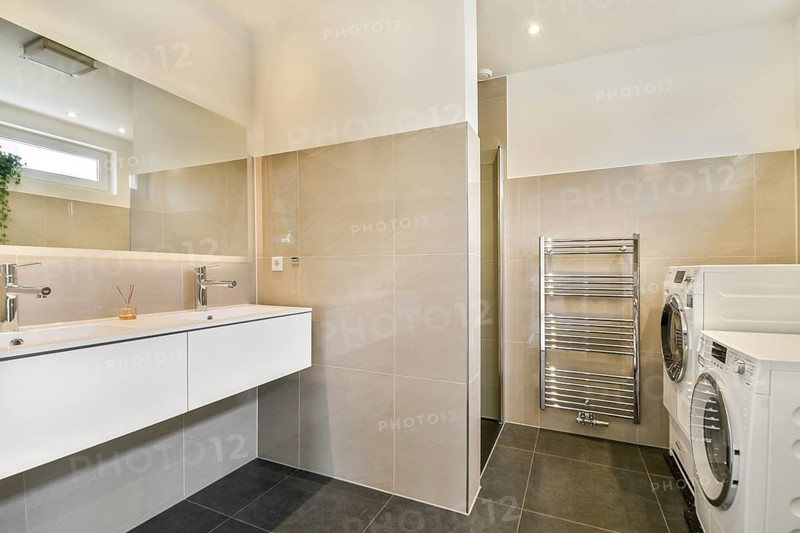 A contemporary bathroom interior featuring a sleek double sink vanity, large mirror, and tiled walls. A modern washer and dryer are neatly integrated into the design