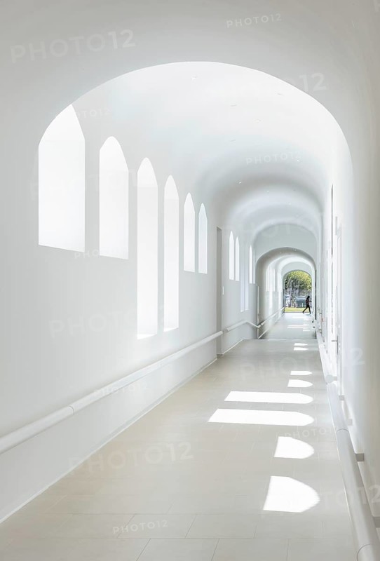 Sunlit corridor of hospital with arched windows creating playful light patterns. The minimalist white design adds a serene atmosphere, inviting exploration and contemplation