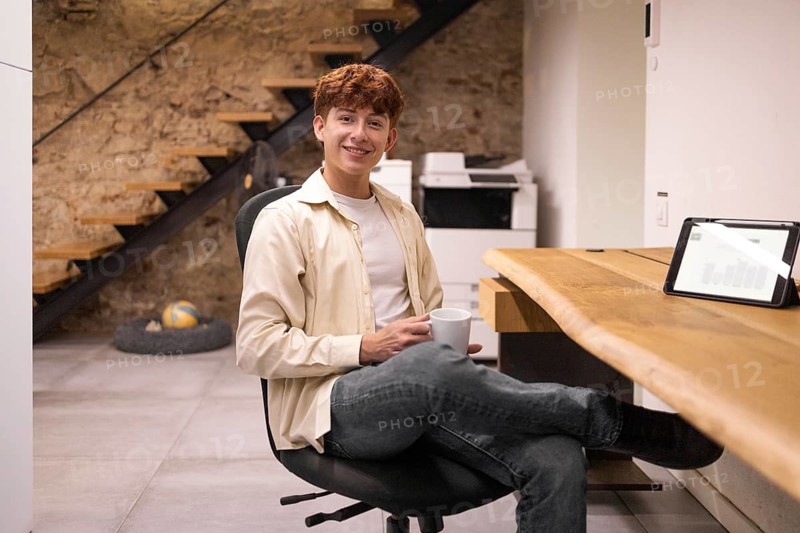 A young man sits casually in a modern coworking space, holding a coffee cup. A tablet displays charts nearby, creating a relaxed work environment