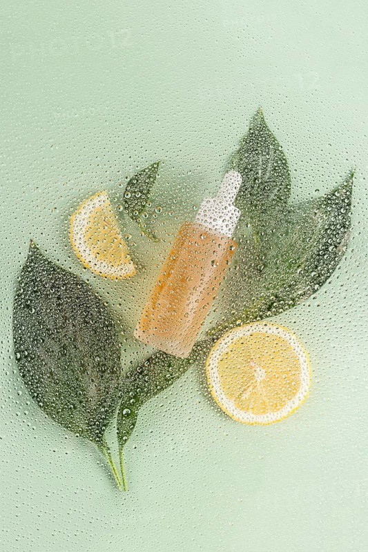 An eye catching display of a skincare cosmetic surrounded by fresh green leaves and lemon slices. Water droplets create a refreshing and natural appeal