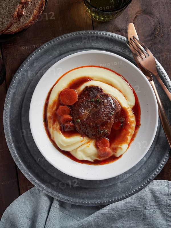 Dish with braised pork cheeks with carrots and gravy over mashed potatoes. Spanish cuisine. View from above, plate on wooden table, golden cutlery and under metallic plate