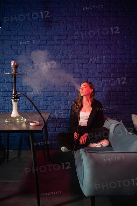 A woman is sitting on a sofa, calmly enjoying a hookah with smoke swirling in a cozy lounge. She is surrounded by cushions and a blue brick wall background