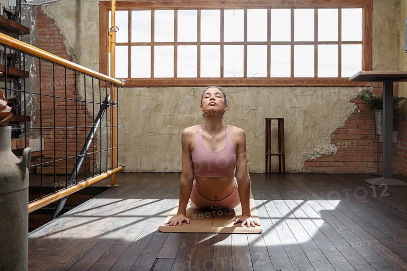 A woman performs yoga in a well lit room with natural sunlight streaming through large windows. The rustic environment adds to the tranquil atmosphere