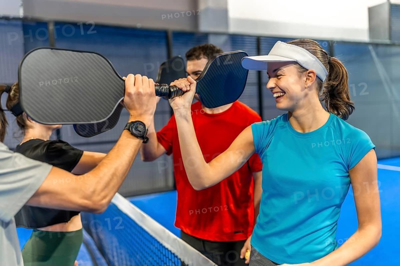 Group of diverse friends dressed in athletic apparel high fiving paddles over the net, celebrating after a successful pickleball match on an indoor court while smiling