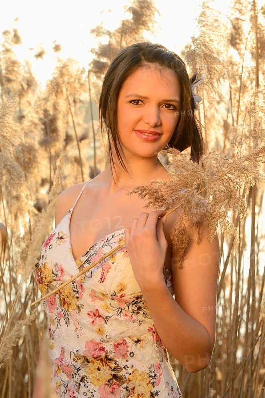 A woman in a floral dress holding a plant and smiling amongst reeds in the serene evening light, Bavaria