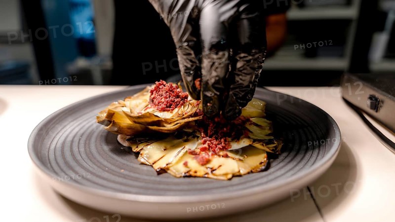 A chef expertly prepares a gourmet dish, showcasing finely chopped ingredients and culinary artistry on a dark plate. The focus is on detail and quality in plating
