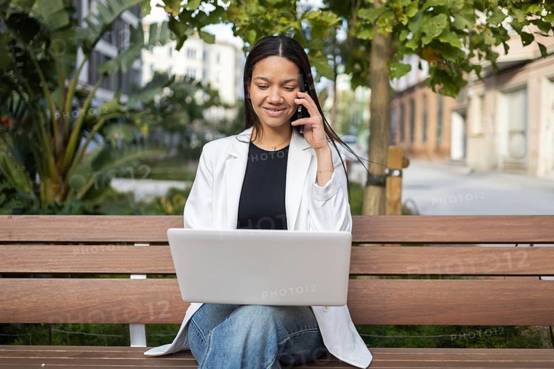 A professional woman in casual attire sits on a bench, working on her laptop while talking on the phone. The outdoor setting blends business with leisure