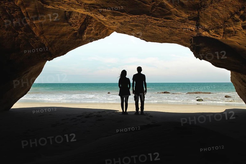 A couple stands inside a cave at Mancora Beach, Peru, gazing at the ocean. The cave's entrance frames the blue sea and sky, creating a stunning and romantic tropical landscape