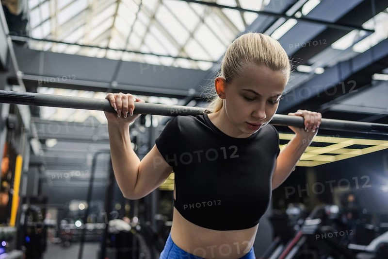 A focused blonde woman lifting a barbell in a contemporary gym. She wears a black crop top and blue leggings. Sunlight streams from a glass ceiling, highlighting the modern interior