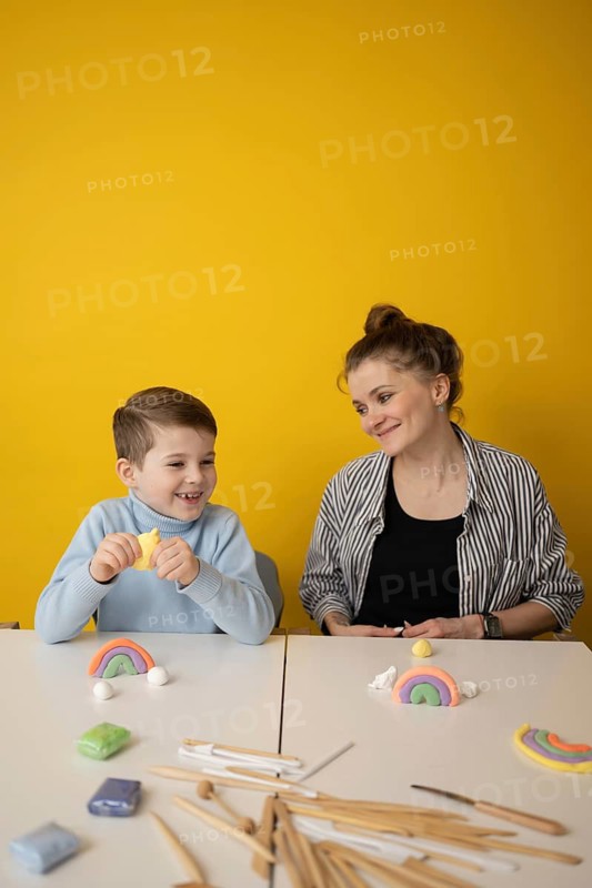 A child and a woman engage in a fun craft session, using colorful clay to create rainbows. The yellow background adds warmth to the creative atmosphere