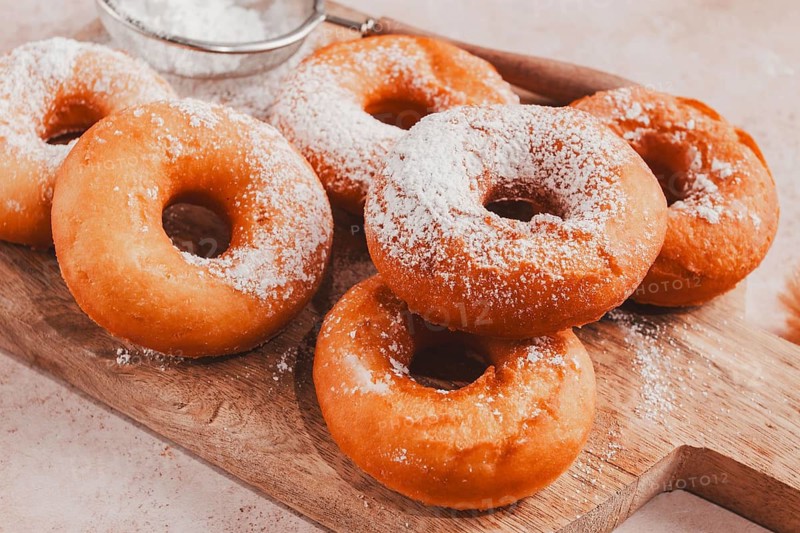 A wooden board holds several freshly made donuts. The donuts are round and have powdered sugar on top. A small sieve is nearby for additional sugar