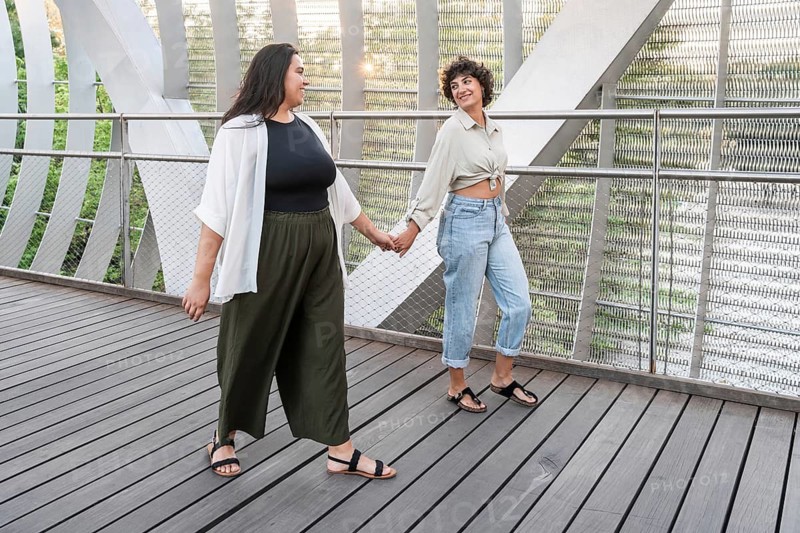 A joyful lesbian couple walks hand in hand on a modern bridge, with metal and wood elements, surrounded by nature, symbolizing love, freedom, and diversity