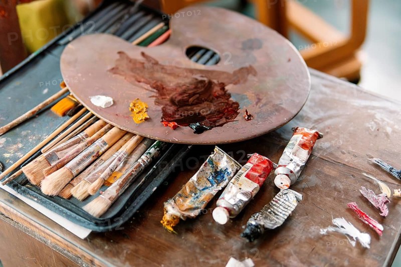 A wooden palette with mixed colors rests on a case of paintbrushes, with used paint tubes nearby. The scene highlights the artistic process and the tools of a dedicated painter