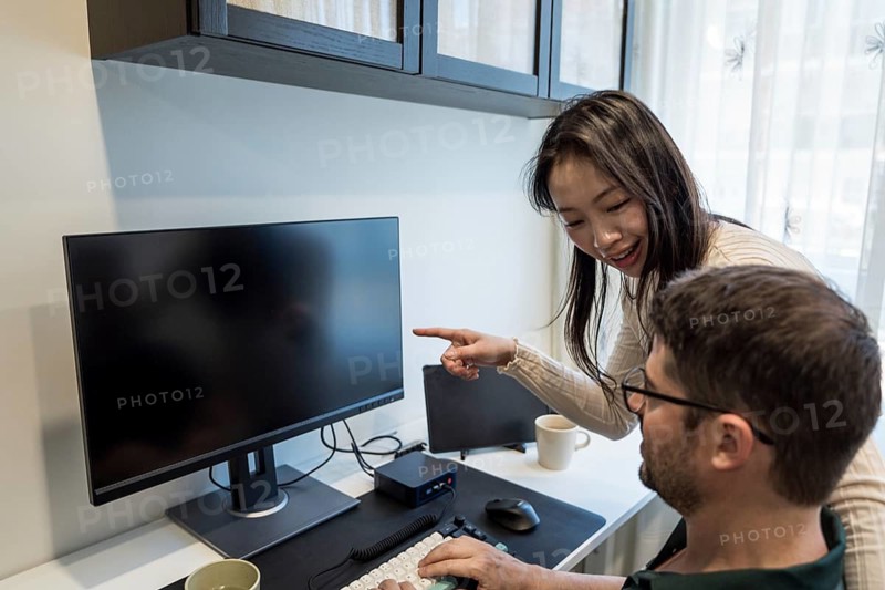 A diverse couple is seen engaging in a collaborative project at home, with the woman pointing to the computer screen, creating an environment of teamwork and camaraderie
