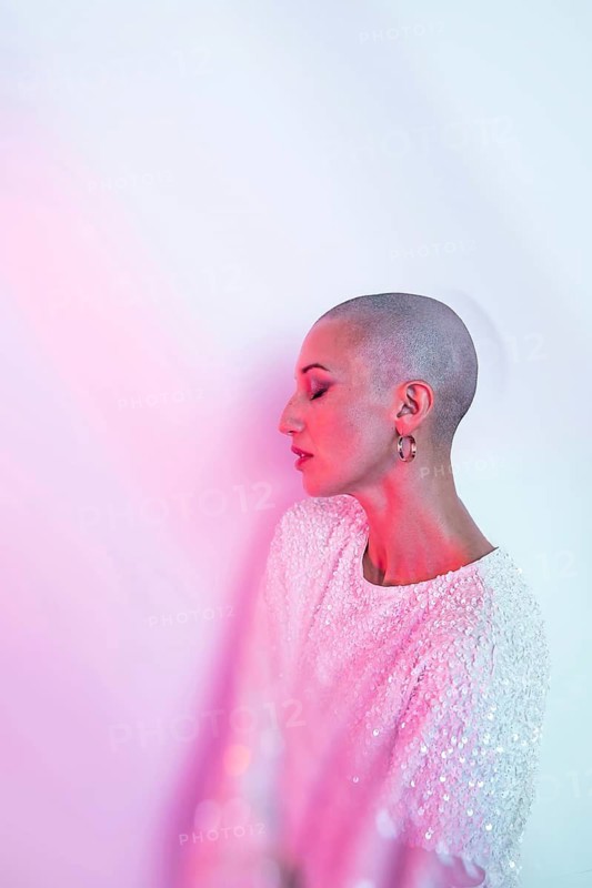 A middle-aged woman with a shaved head poses elegantly, wearing a sparkling top. The soft pink lighting adds a serene and artistic touch to the portrait