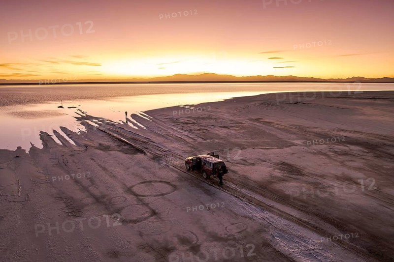 A stunning sunset casts a warm glow over Salinas Grandes, Salta, Argentina. An off-road vehicle is parked on the expansive salt flats, offering an adventurous escape in this breathtaking landscape