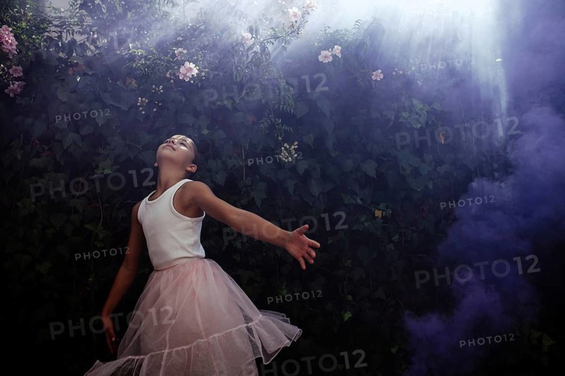 A teenage girl in a pink tulle skirt dances gracefully under the foliage, basking in the sunshine and surrounded by mist and light smoke. The scene radiates joy, freedom, and connection with nature