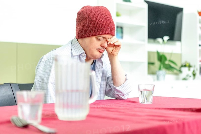 A man in a cozy red beanie and casual shirt sits at a table in a mental health residence, appearing thoughtful and reflective. A water jug and glass are nearby, creating a calm atmosphere