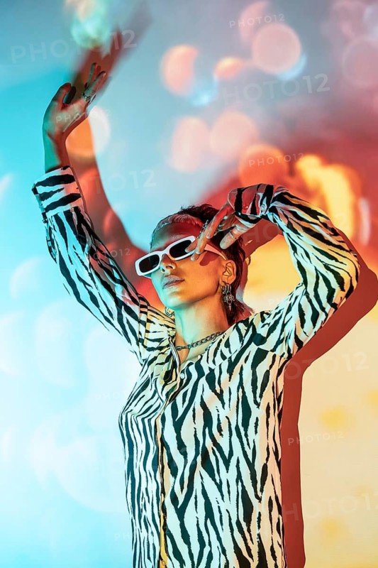 Young trendy woman poses confidently, wearing bold zebra print and white sunglasses, with colorful bokeh background creating a vivid, dynamic atmosphere in modern fashion