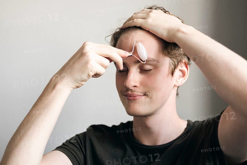 A young male rests at home while using a facial roller to massage his forehead. He appears relaxed, promoting skincare and wellness