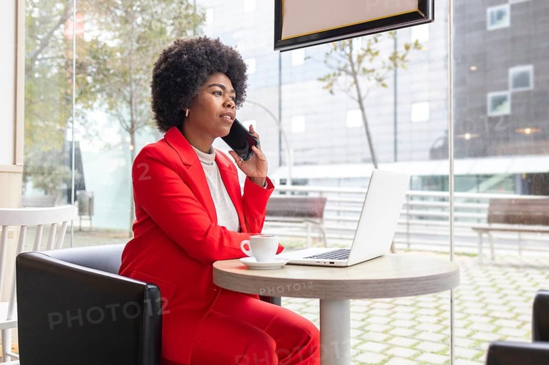 A professional woman in a red suit sits in a modern cafe, talking on her phone while working on a laptop. This image captures a dynamic urban lifestyle