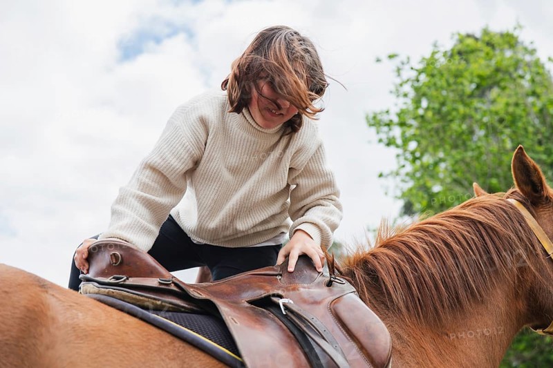 A young girl, wearing a cozy sweater, enjoys a sunny day on a farm as she prepares to ride a beautiful brown horse. The sky is bright, enhancing the vibrant scene