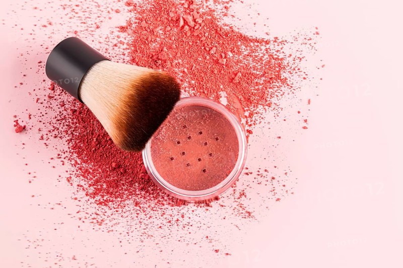 A compact blush sits on a pastel pink surface with a makeup brush. The soft bristles are ideal for applying the blush powder, creating a natural and radiant finish