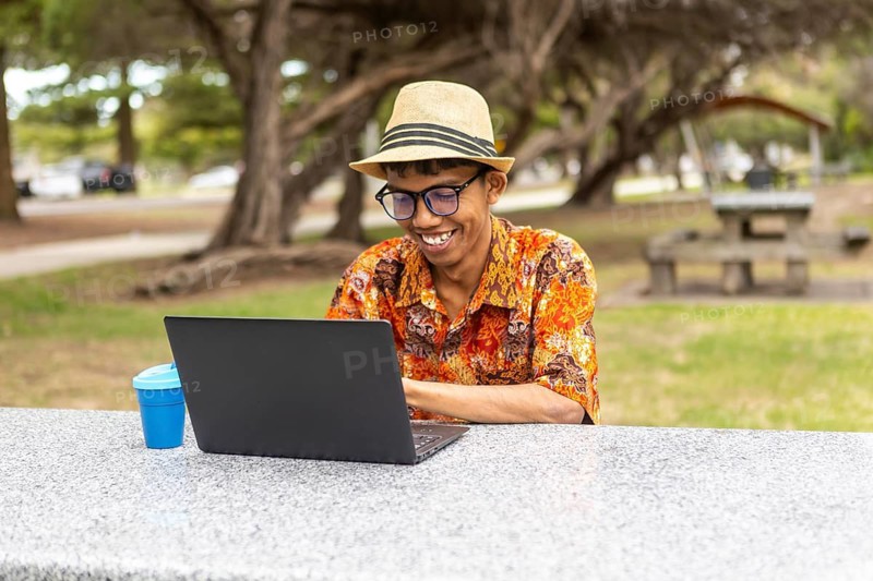 An indonesian man wearing vibrant batik clothing and a hat uses a laptop outdoors in a park. The setting combines Indonesian culture with an Australian backdrop