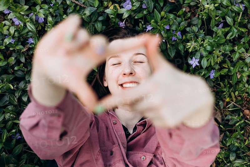 Joyful young woman lying on a bed of blooming periwinkle flowers, smiling and playfully framing the scene with her hands in a natural green setting