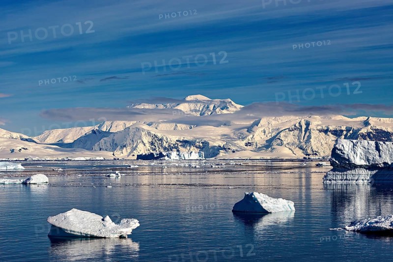 Isolated icebergs in still water in front of a snow-covered mountain backdrop under a slightly cloudy sky, icebergs in the landscape of the Southern Ocean in Antarctica
