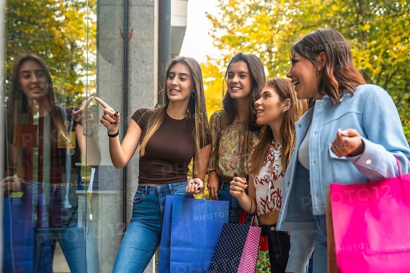 Group of happy young women with shopping bags pointing, smiling, and looking at a storefront display while enjoying retail activity outdoors in the city