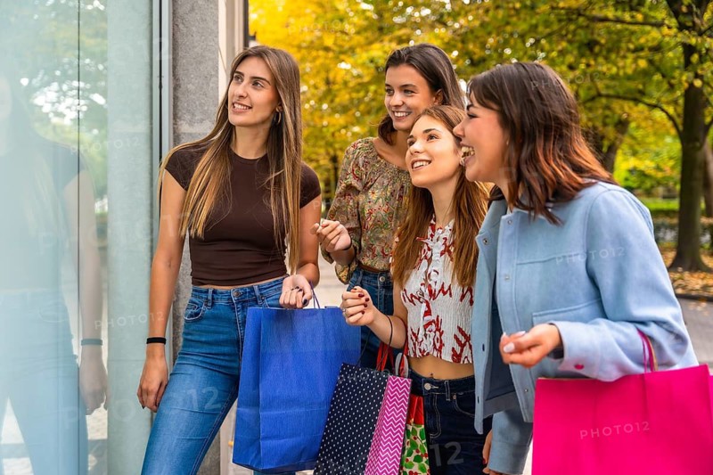 Group of happy women friends walking together on a city street, looking at a shop window with colorful shopping bags, celebrating friendship and consumerism in autumn