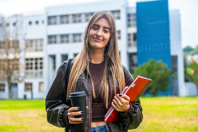 Young female student smiling and standing on a university campus, holding a takeaway coffee cup and red notebooks, ready for her classes and educational journey