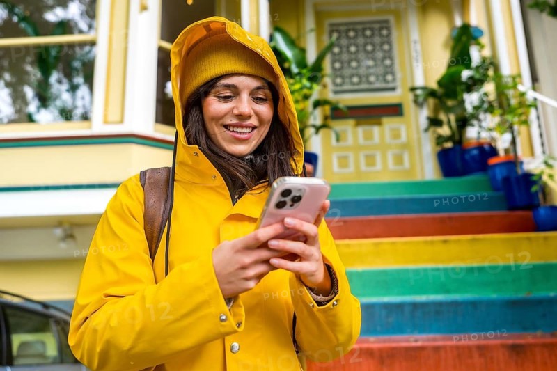 Woman in vibrant yellow raincoat and beanie smiling at smartphone on san francisco street, colorful rainbow stairs and victorian house in background, urban travel and connectivity