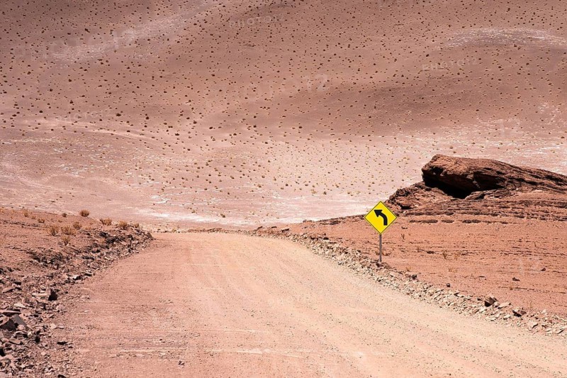 A dusty, winding road marked by a yellow road sign indicating a curve, located in the arid, desert region of La Puna, Argentina. This surreal landscape showcases a vast, barren terrain under a clear sky