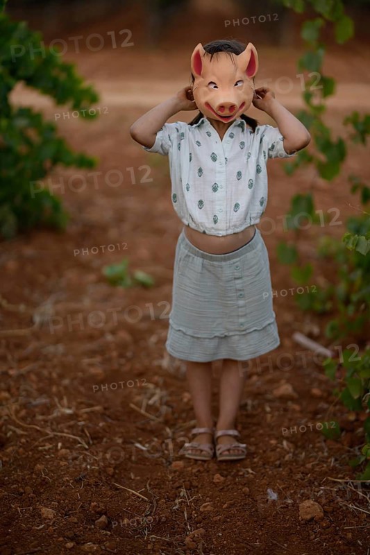 A young child stands in a vineyard, playfully holding a pig mask over their head. The scene highlights creativity and fun in a natural, sunny outdoor setting