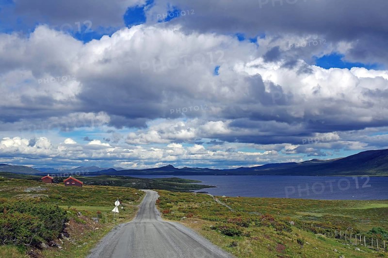A rural road passes by a lake surrounded by mountains and a dramatic sky, Fjell, Jotunheimvegen, Valdresflyra, Norway