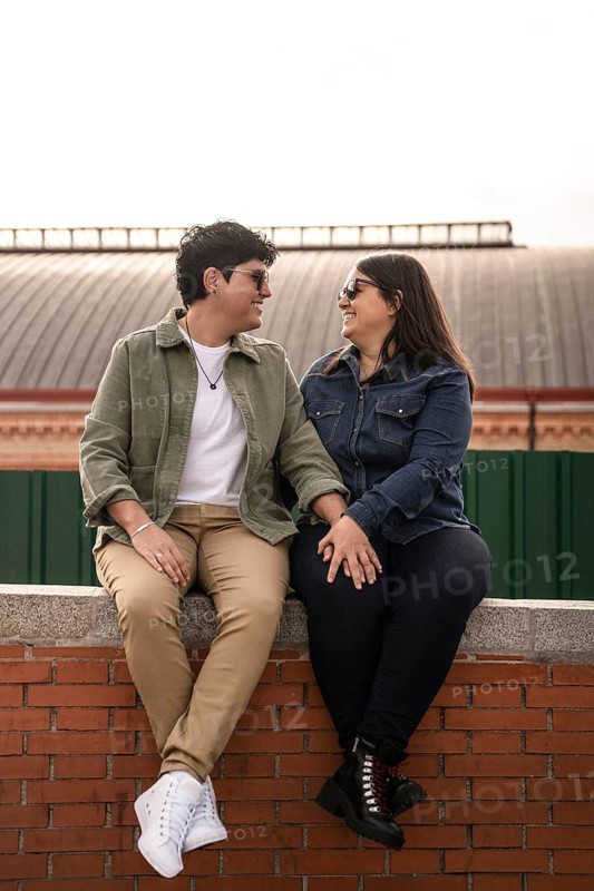 A joyful lesbian couple sits on a brick wall, holding hands and sharing a tender moment. Both women are smiling and gazing at each other with affection