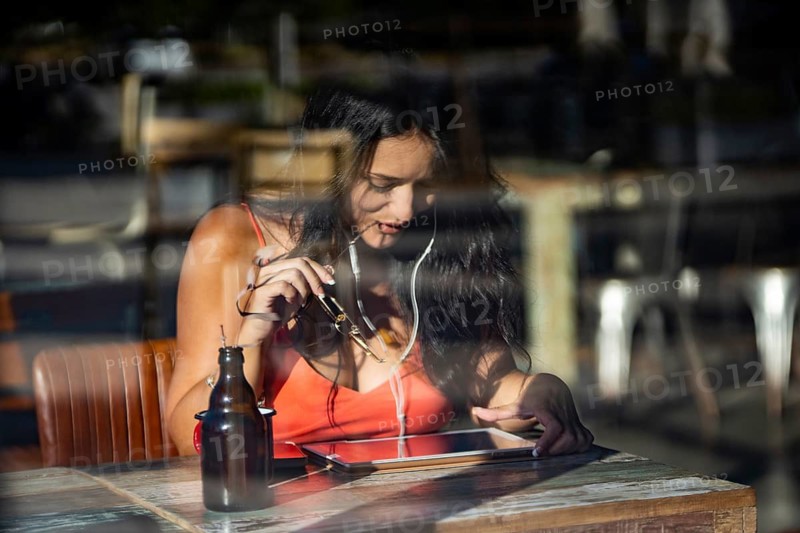A woman sits in a cafe, absorbed in her tablet and wearing headphones. Sunlight streams in, casting reflections on the window. A drink bottle rests on the wooden table