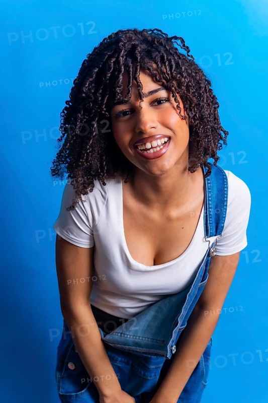 Portrait of a cheerful young woman with curly hair, wearing denim overalls and a white t shirt, smiling against a vibrant blue backdrop