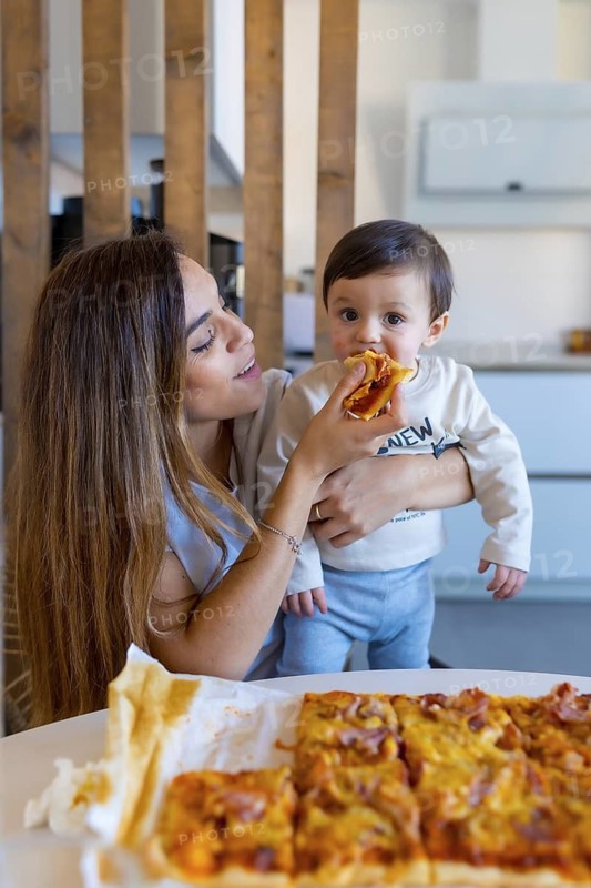 Mom holding her baby and feeding him a slice of pizza, sitting at the kitchen table with a whole pizza ready to be eaten