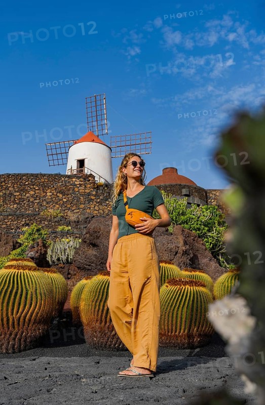 A woman in stylish, casual attire admires a traditional windmill in Lanzarote, set against a backdrop of cacti and blue sky during her holiday visit to the Canary Islands, Spain