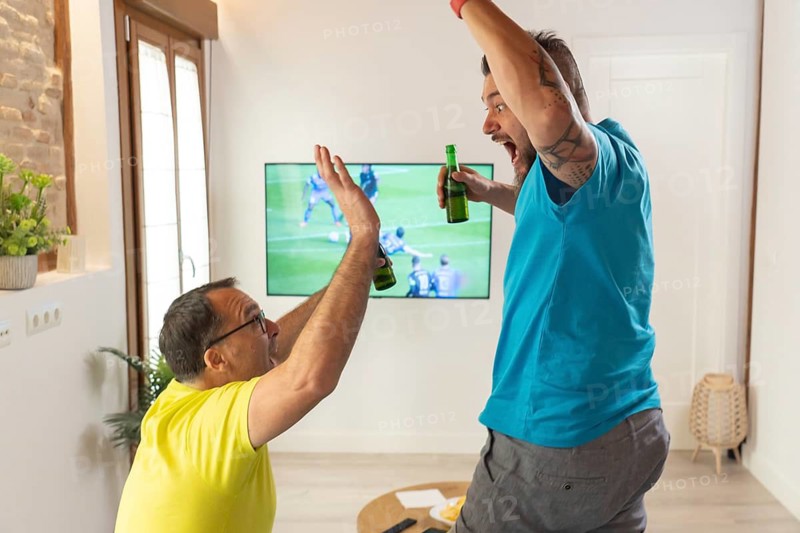 Two friends celebrating a goal with a high five while watching a soccer match on TV at home. Excitement, joy, beer and friendship indoors