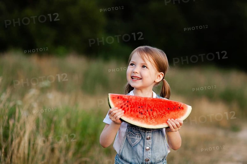 A cheerful child holding a slice of watermelon, enjoying the warm summer day outdoors in a field. The bright red fruit and the child's joy capture the essence of summer fun