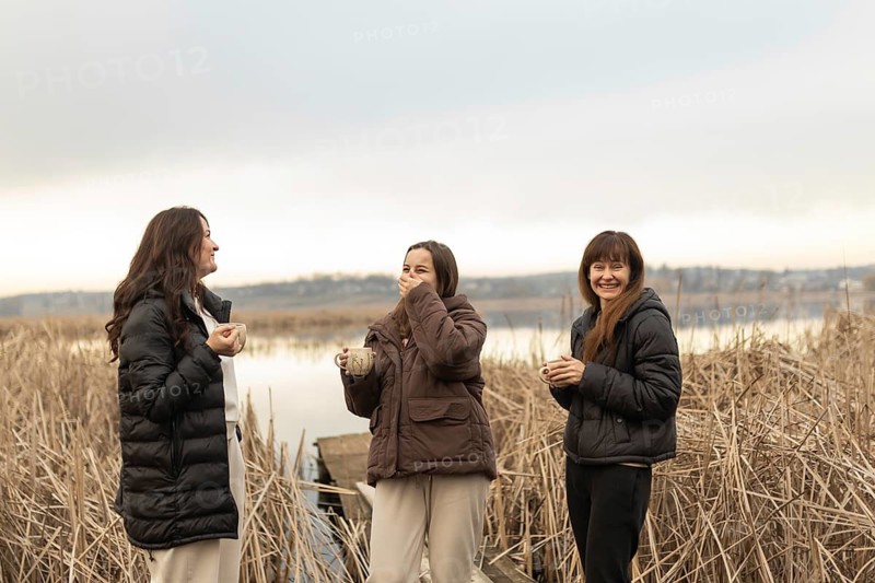 Three female friends share laughter and conversation while holding warm drinks, standing against a natural lakeside backdrop with dry reeds. They enjoy a cozy get-together in cool weather attire