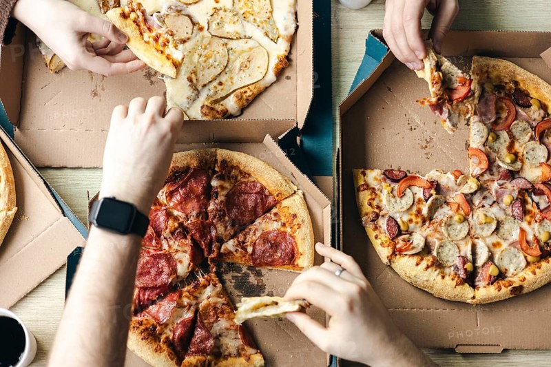 Top view of a group of unrecognizable friends gathers around the table, sharing slices of various pizzas from cardboard boxes. The image captures a casual and friendly meal setting