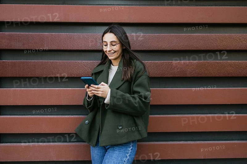 A woman in a green coat is smiling while using her smartphone She stands against a decorative striped wall, creating a casual yet stylish urban scene