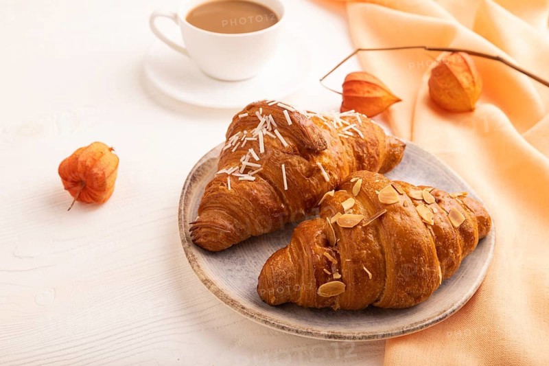 Croissant on blue plate on white wooden background and orange linen textile, cup of coffee, side view, close up