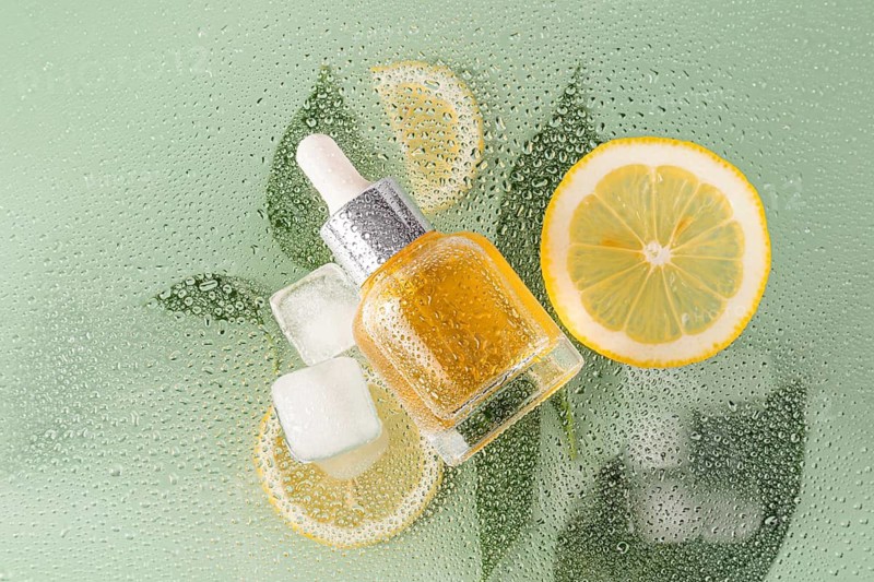 A cosmetic of citrus essence lies on a fresh, dewy surface with lemon slices and ice cubes. The backdrop highlights the refreshing and invigorating mood