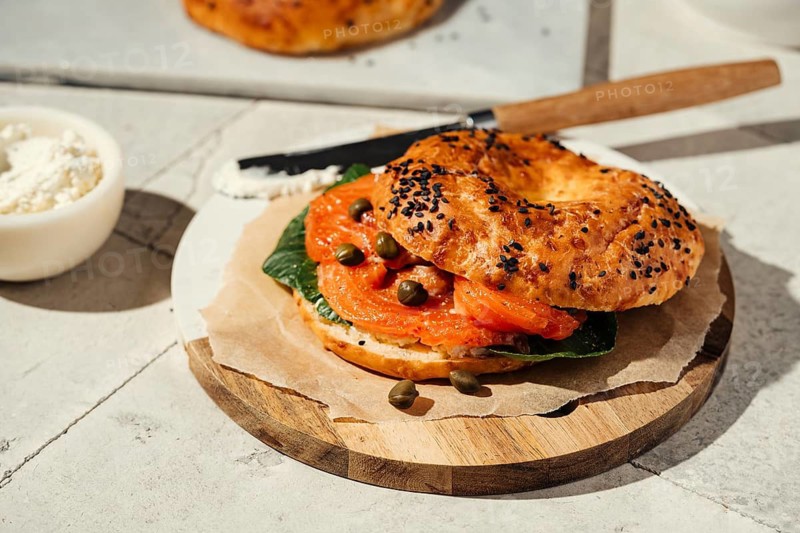 A freshly baked sesame bagel filled with smoked salmon, capers, and greens on a wooden plate. Light cream cheese is spread with a knife, adding a creamy texture